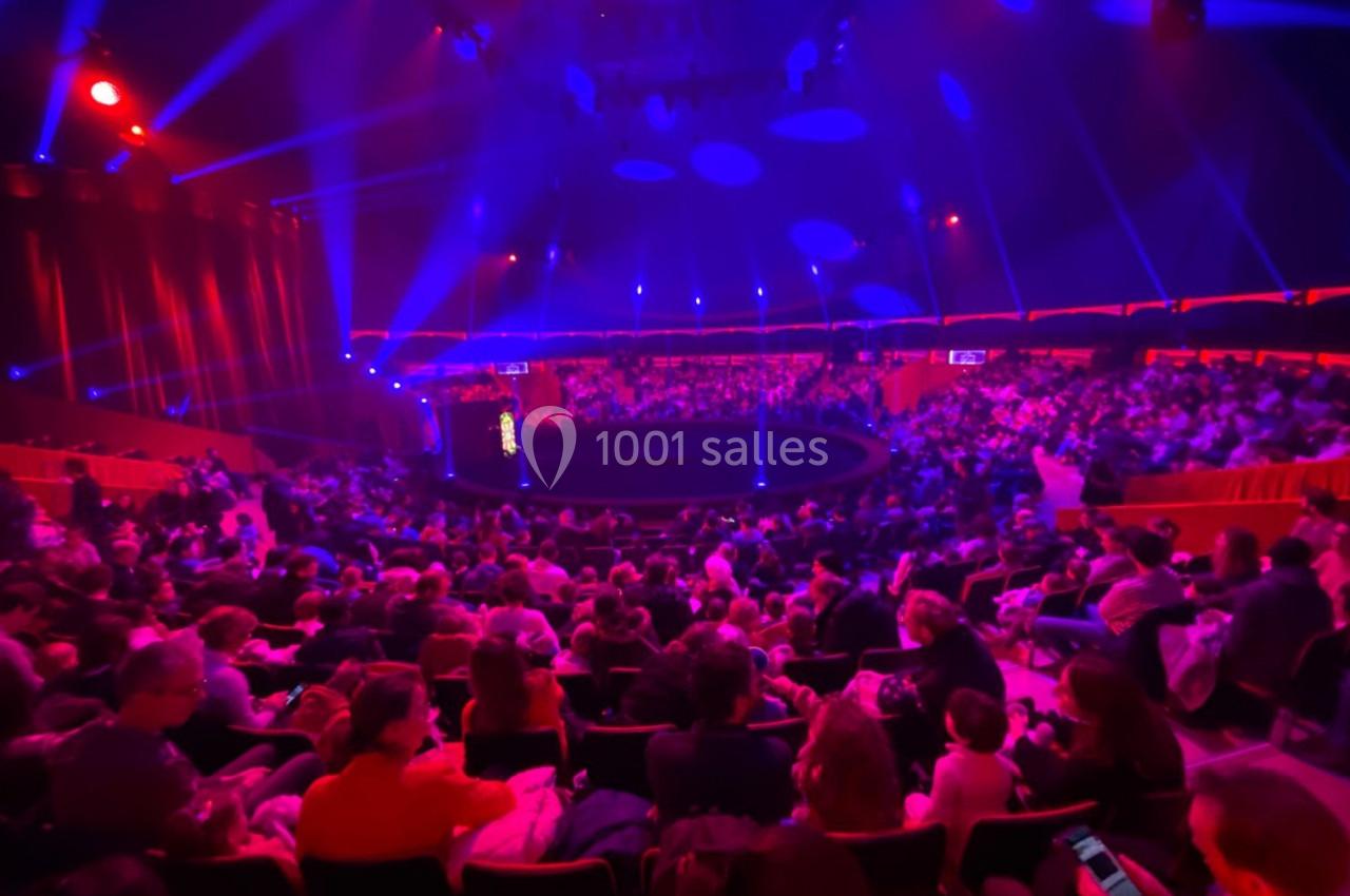 Public assis dans une salle de spectacle circulaire, éclairée par des lumières rouges et bleues.