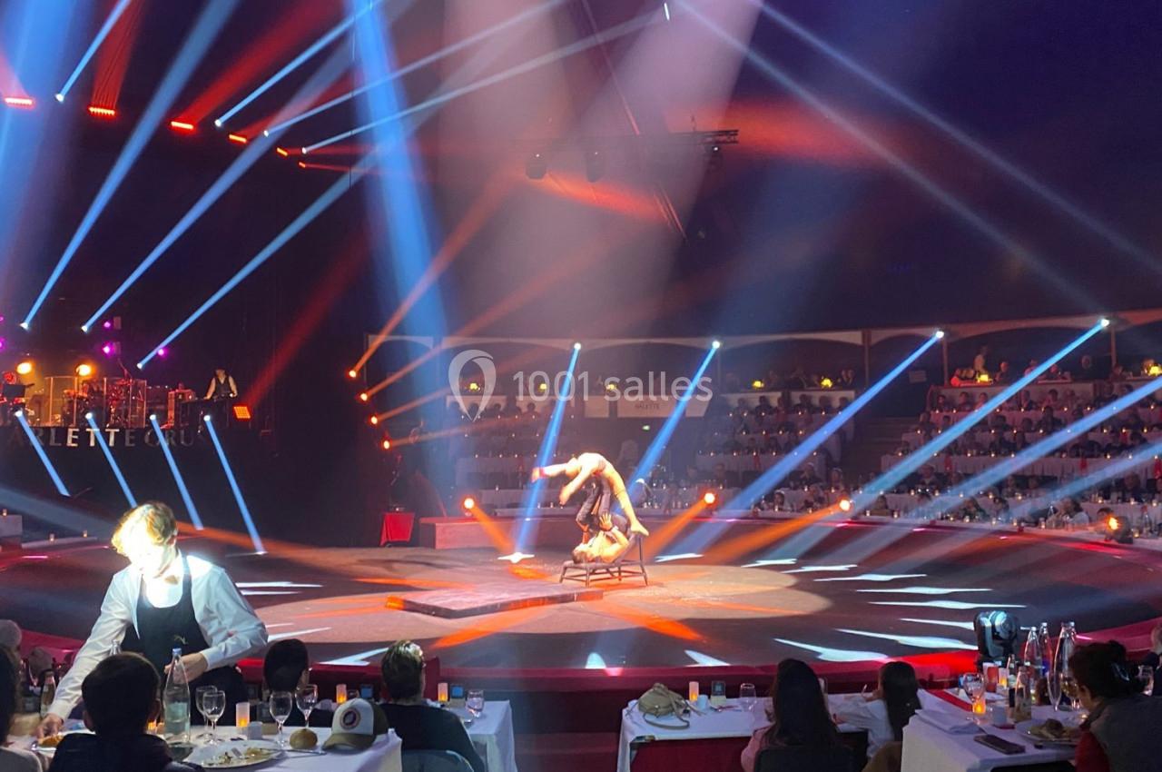 Un artiste réalise une performance acrobatique sur une chaise au centre d'une scène éclairée, devant un public assis.