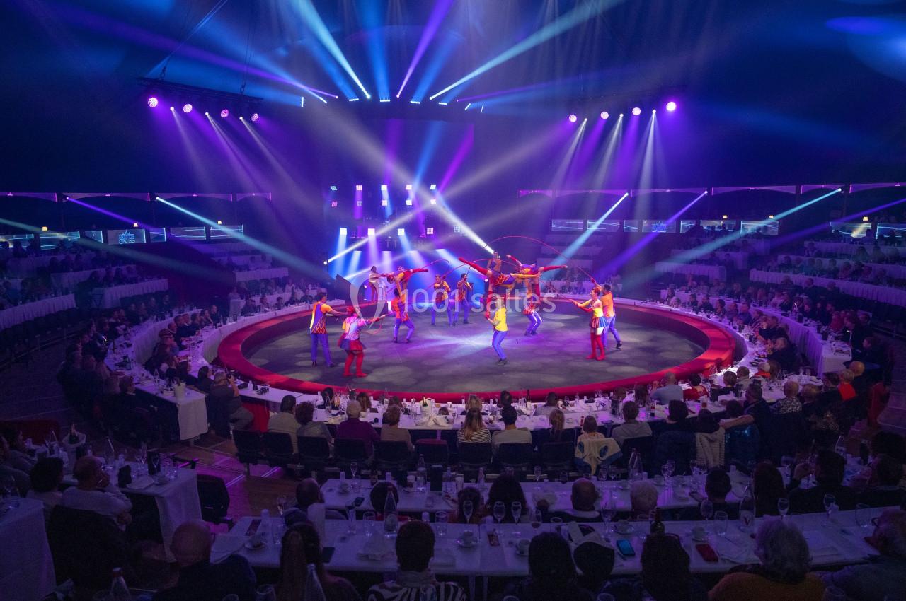 Des artistes exécutent une performance colorée sur une scène circulaire entourée de spectateurs sous des lumières vives.