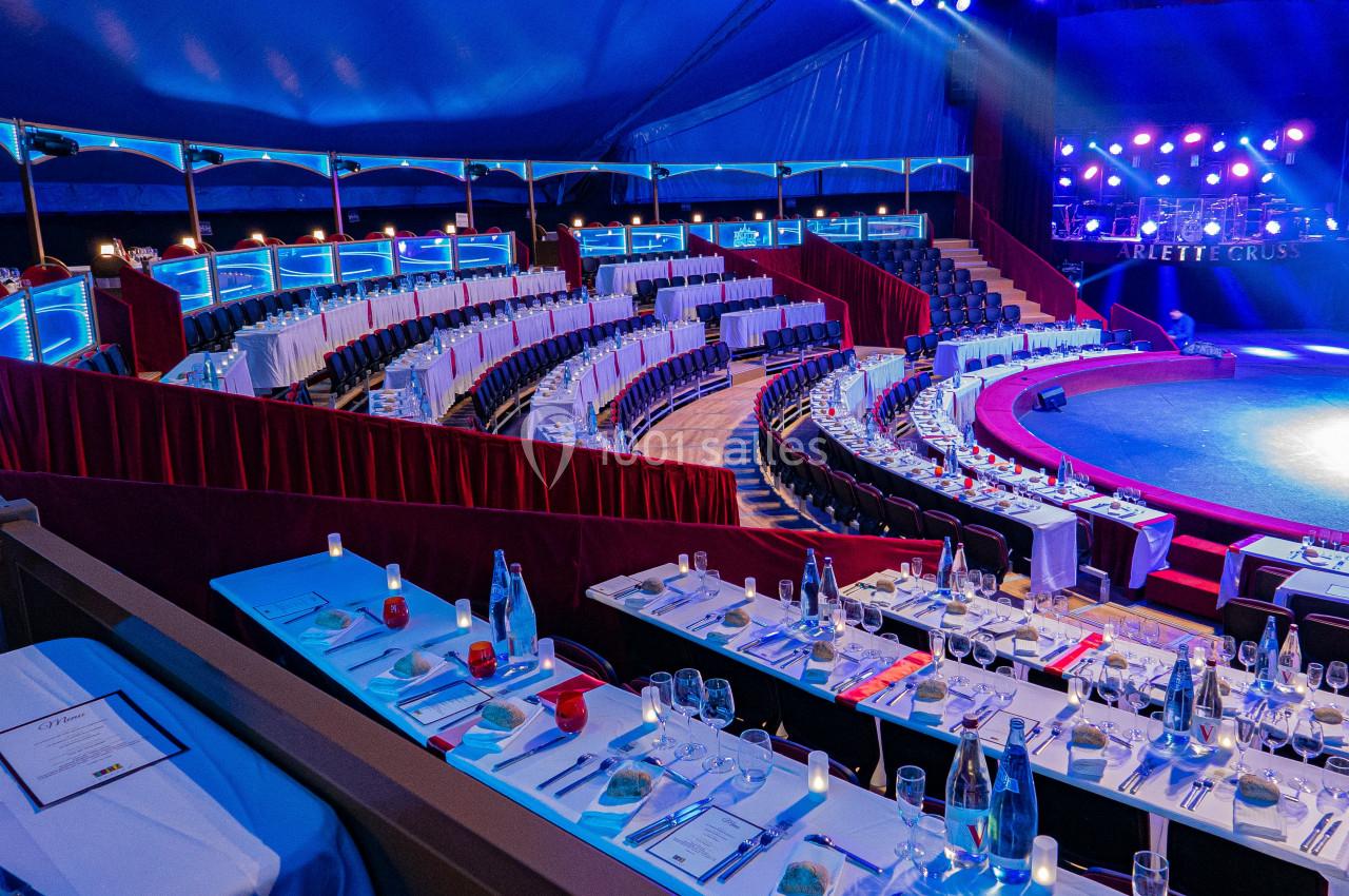 Gradins et tables dressées sous un chapiteau de cirque, éclairés par des lumières colorées.