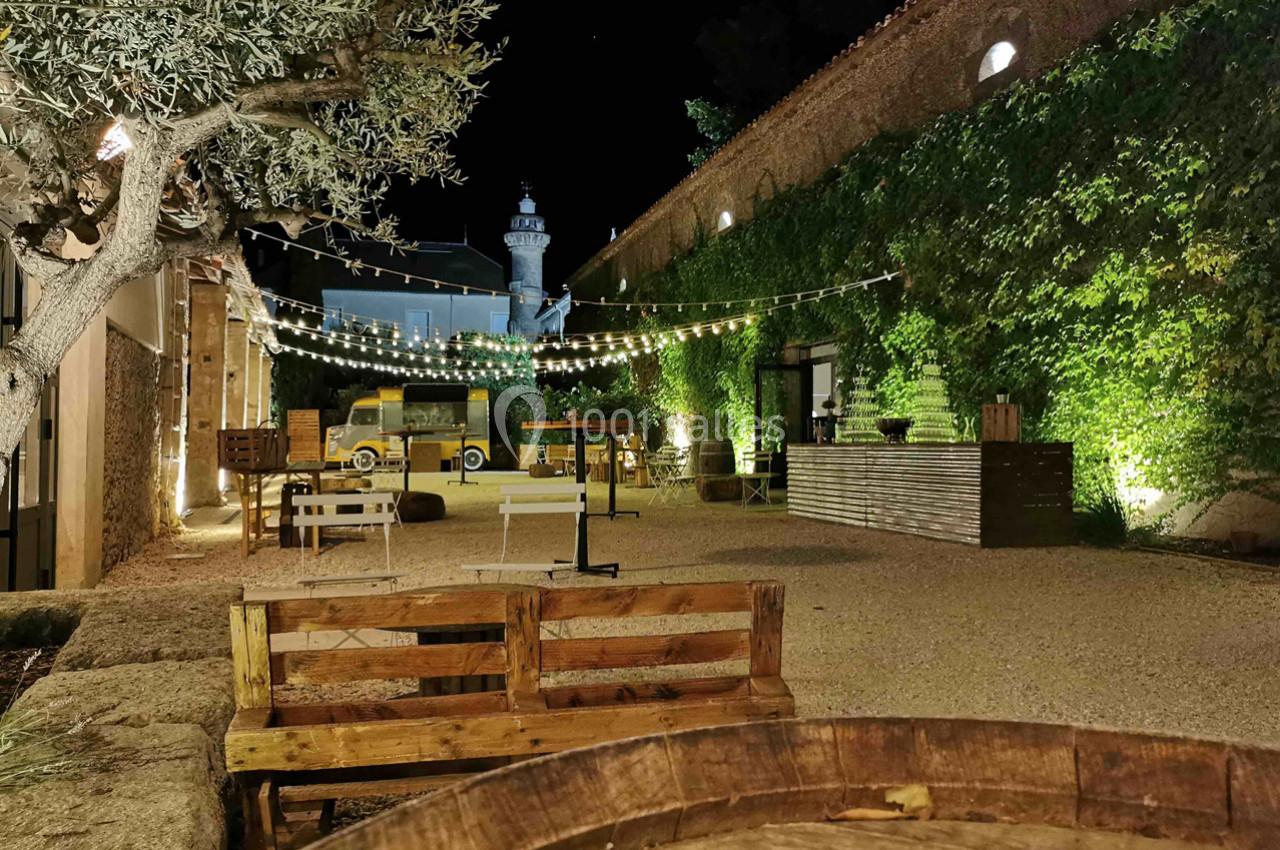 Cour extérieure éclairée la nuit, avec guirlandes lumineuses, bancs en bois, bar et végétation sur les murs.