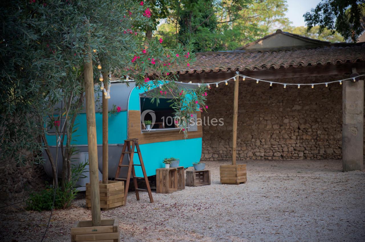 Une caravane turquoise aménagée en stand, entourée de guirlandes lumineuses et de végétation dans une cour en pierre.