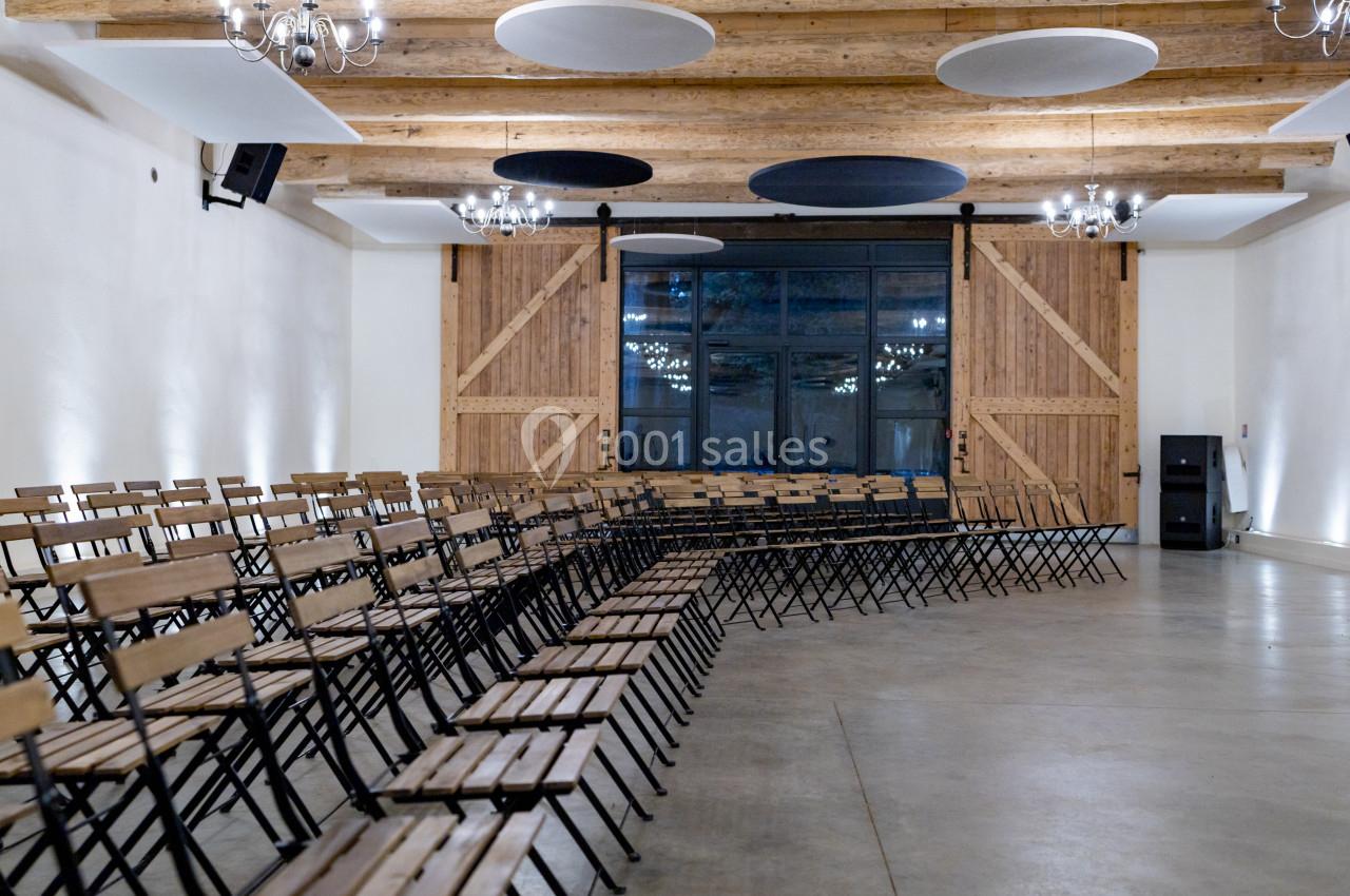 Salle avec des rangées de chaises pliantes en bois, plafond avec poutres apparentes et panneaux acoustiques circulaires.