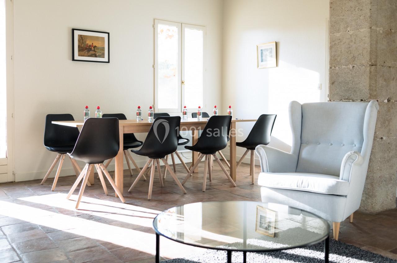 Salle lumineuse avec table en bois entourée de chaises noires, fauteuil gris et table basse en verre.