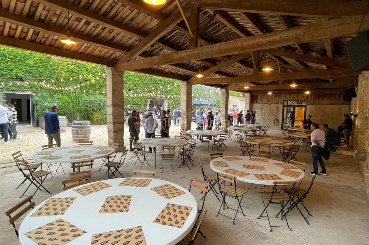 Salle de réception en plein air avec tables rondes dressées, chaises pliantes et invités debout en arrière-plan.