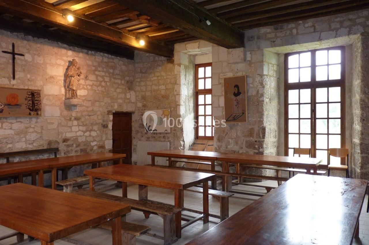 Salle en pierre avec des tables en bois, des chaises alignées et des décorations murales, éclairée par des fenêtres.