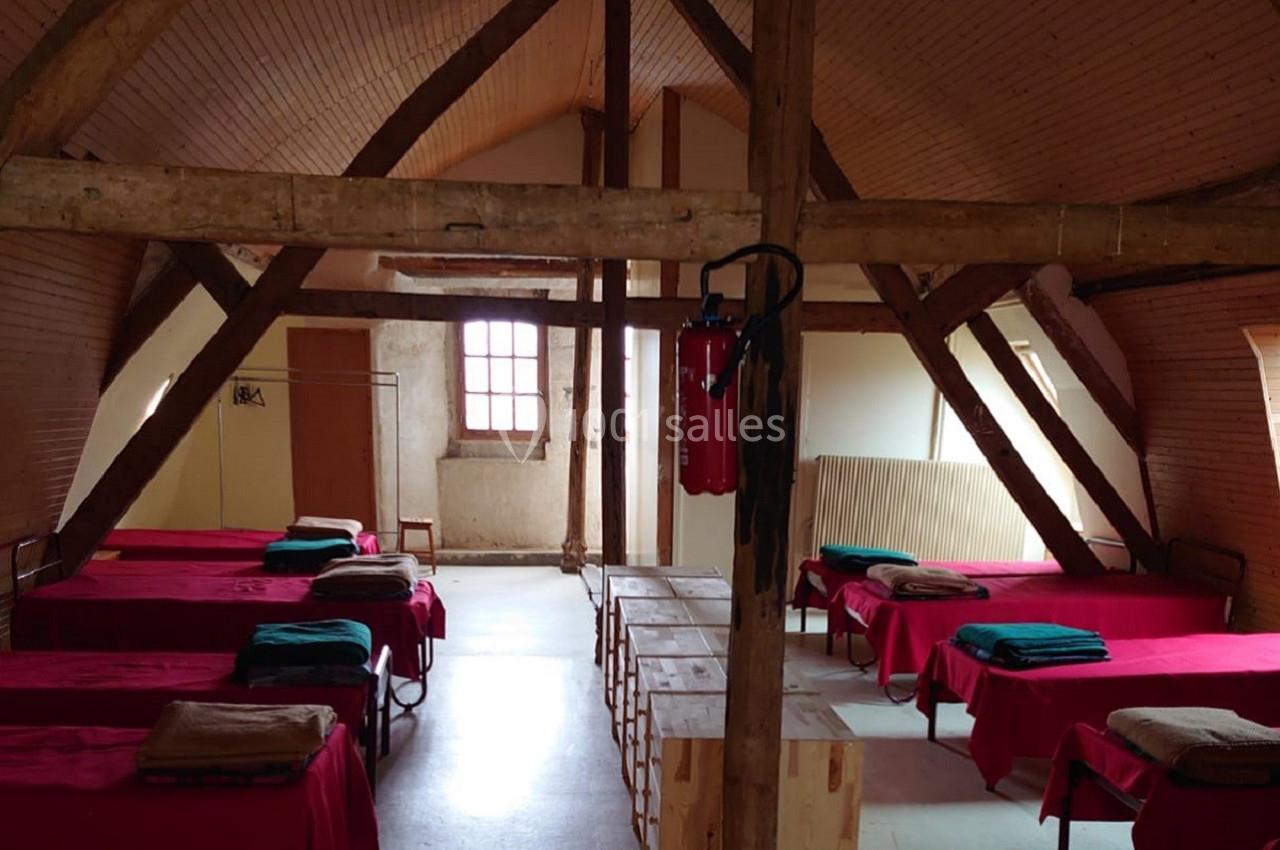 Dortoir avec lits simples alignés, couvertures pliées, poutres apparentes et lumière naturelle provenant d'une fenêtre.