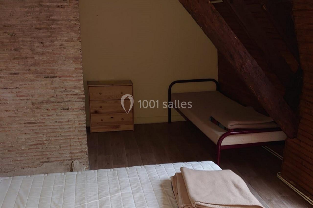 Chambre mansardée avec un lit simple, une commode en bois, un mur en briques et des poutres apparentes.