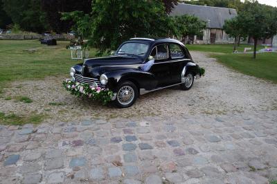 Voiture ancienne noire décorée de fleurs, stationnée sur une allée pavée dans un cadre champêtre.
