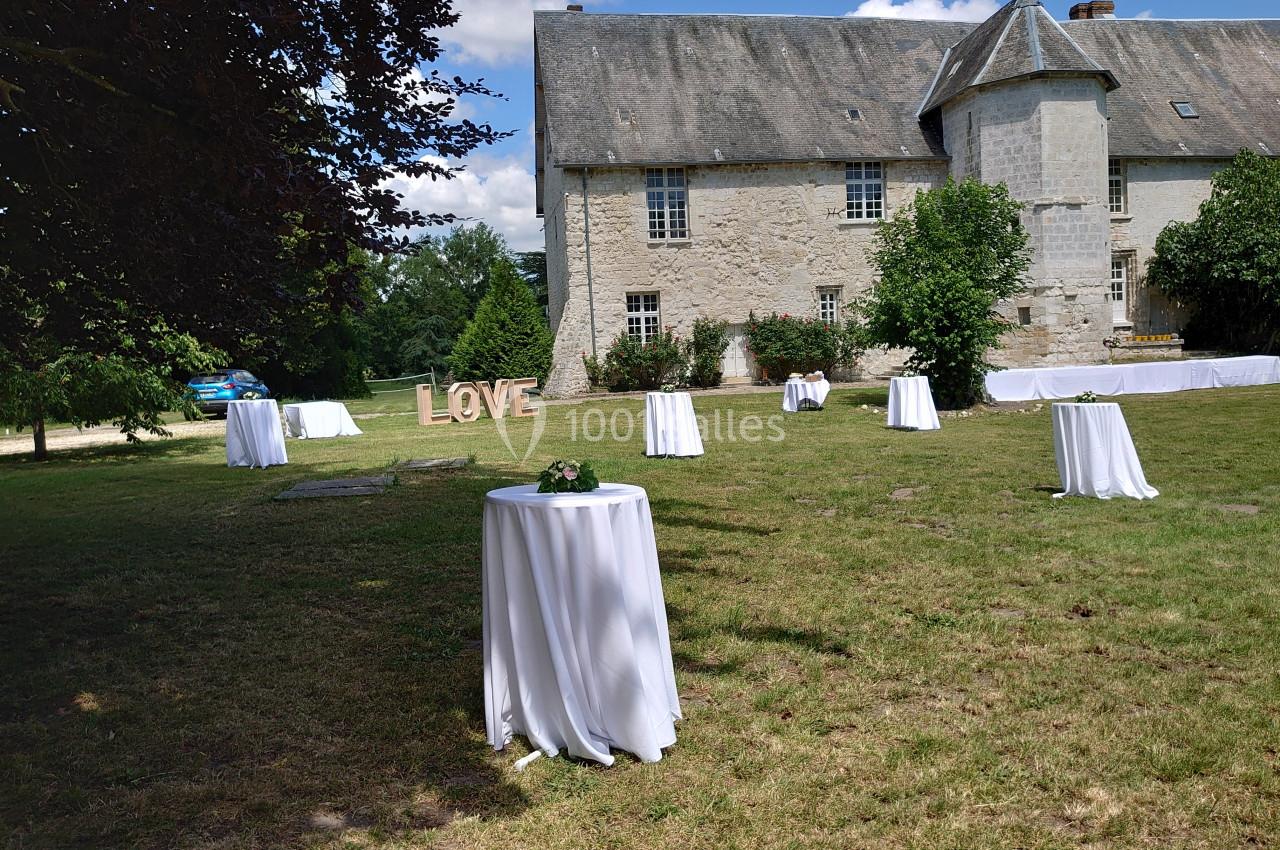 Tables hautes drapées de blanc disposées sur une pelouse devant un bâtiment en pierre, avec des lettres ’LOVE’ en arrière…