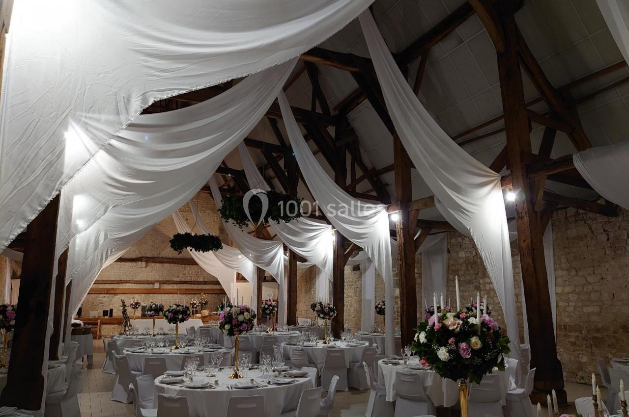 Salle de réception décorée avec des drapés blancs suspendus, tables rondes dressées et ornées de bouquets de fleurs.