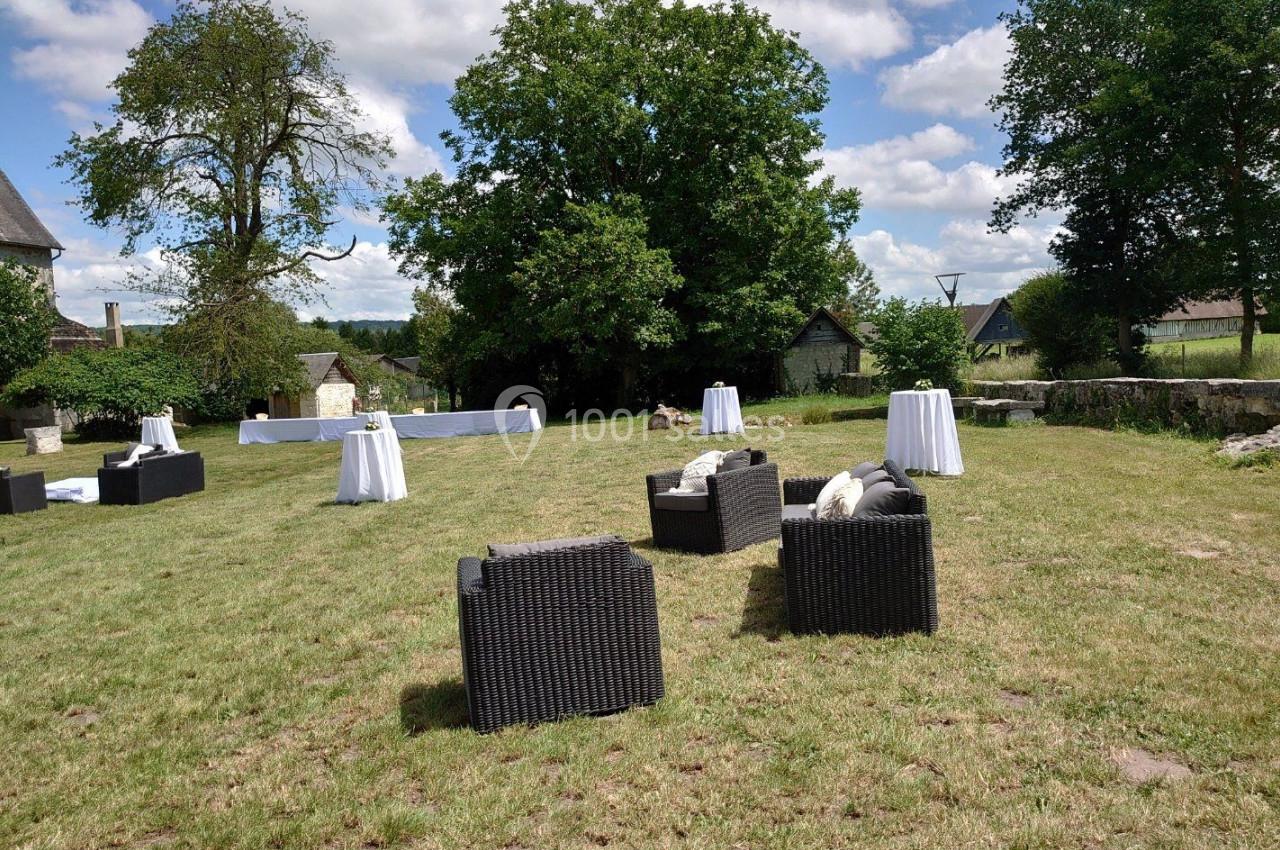 Espace extérieur avec fauteuils en rotin, tables hautes et pelouse, entouré d'arbres et bâtiments en arrière-plan.