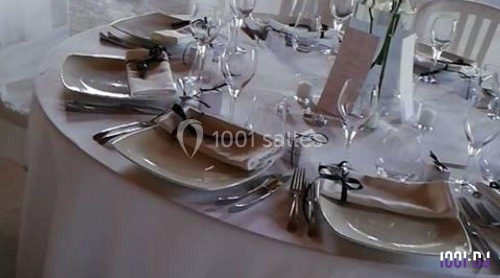 Table ronde dressée avec nappes blanches, assiettes, couverts, verres et serviettes ornées de rubans noirs.