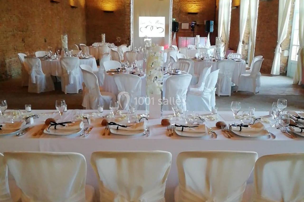 Salle de réception avec tables dressées, nappes blanches, chaises décorées et éclairage naturel à travers de grandes…