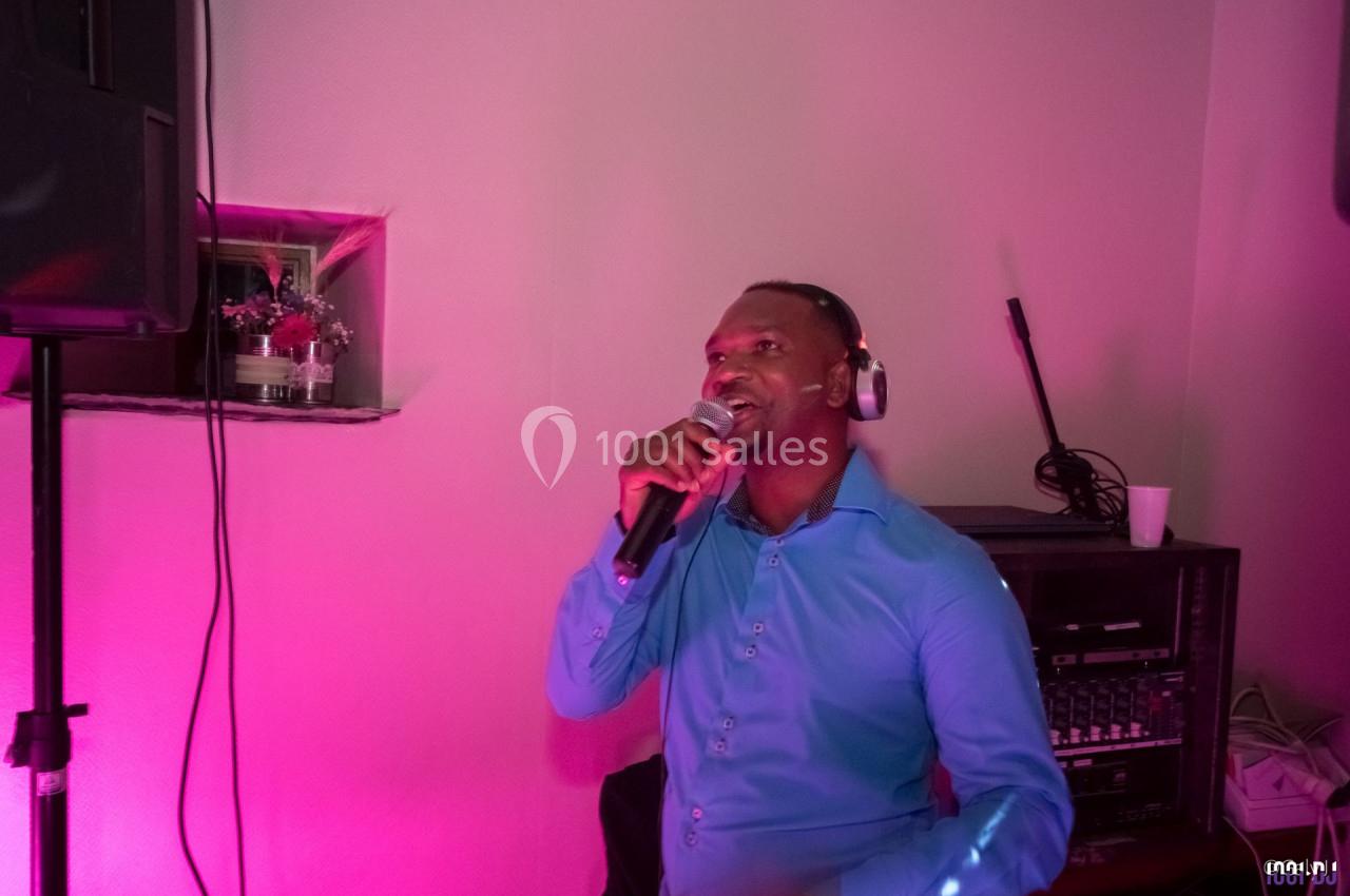 Un homme en chemise bleue chante au micro avec un casque audio, éclairé par une lumière rose dans une pièce.