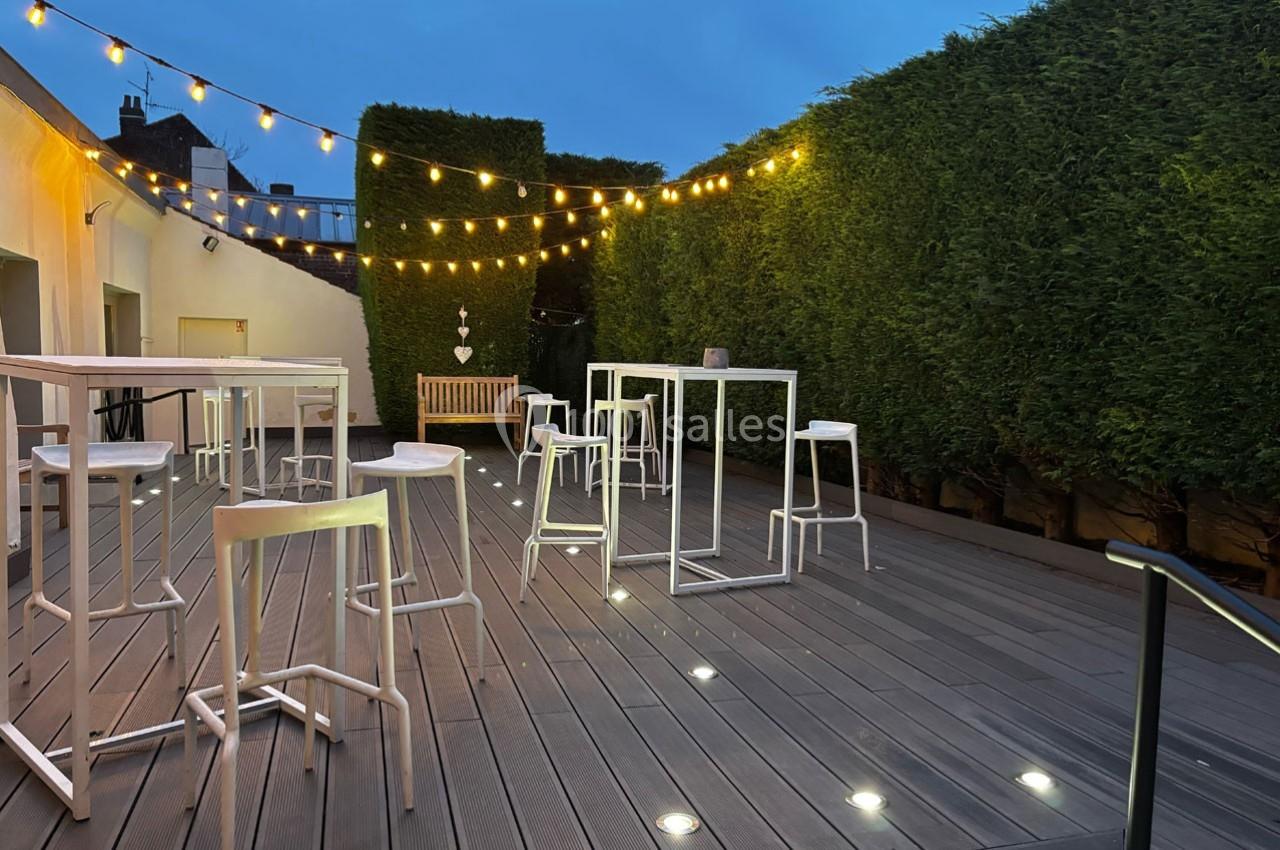 Traiteur Forest-sur-Marque (Nord) - Auberge du Tilleul #15 Terrasse en bois avec tables hautes, tabourets blancs, guirlandes lumineuses et haies taillées en soirée.