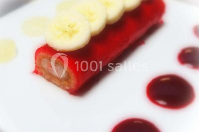 Dessert roulé recouvert de glaçage rouge, garni de rondelles de banane, avec des touches de coulis sur l'assiette blanche.