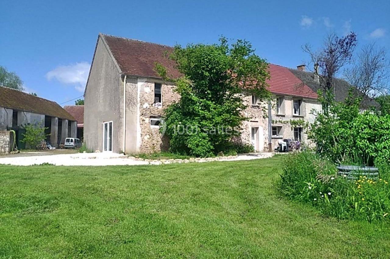 Grande maison en pierre avec toit en tuiles rouges, entourée d'une pelouse et d'arbres sous un ciel bleu.