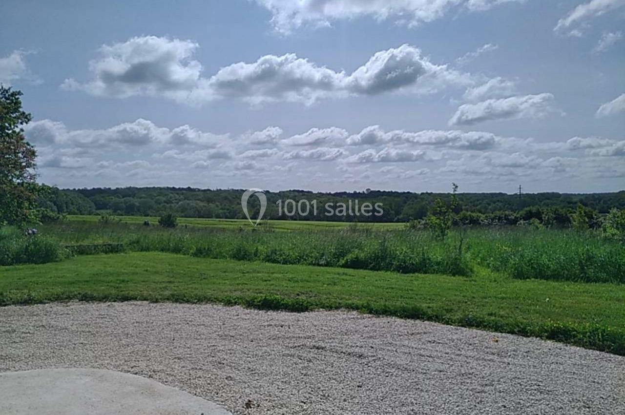 Vue d'un paysage champêtre avec une pelouse, des arbres à l'horizon et un ciel partiellement nuageux.