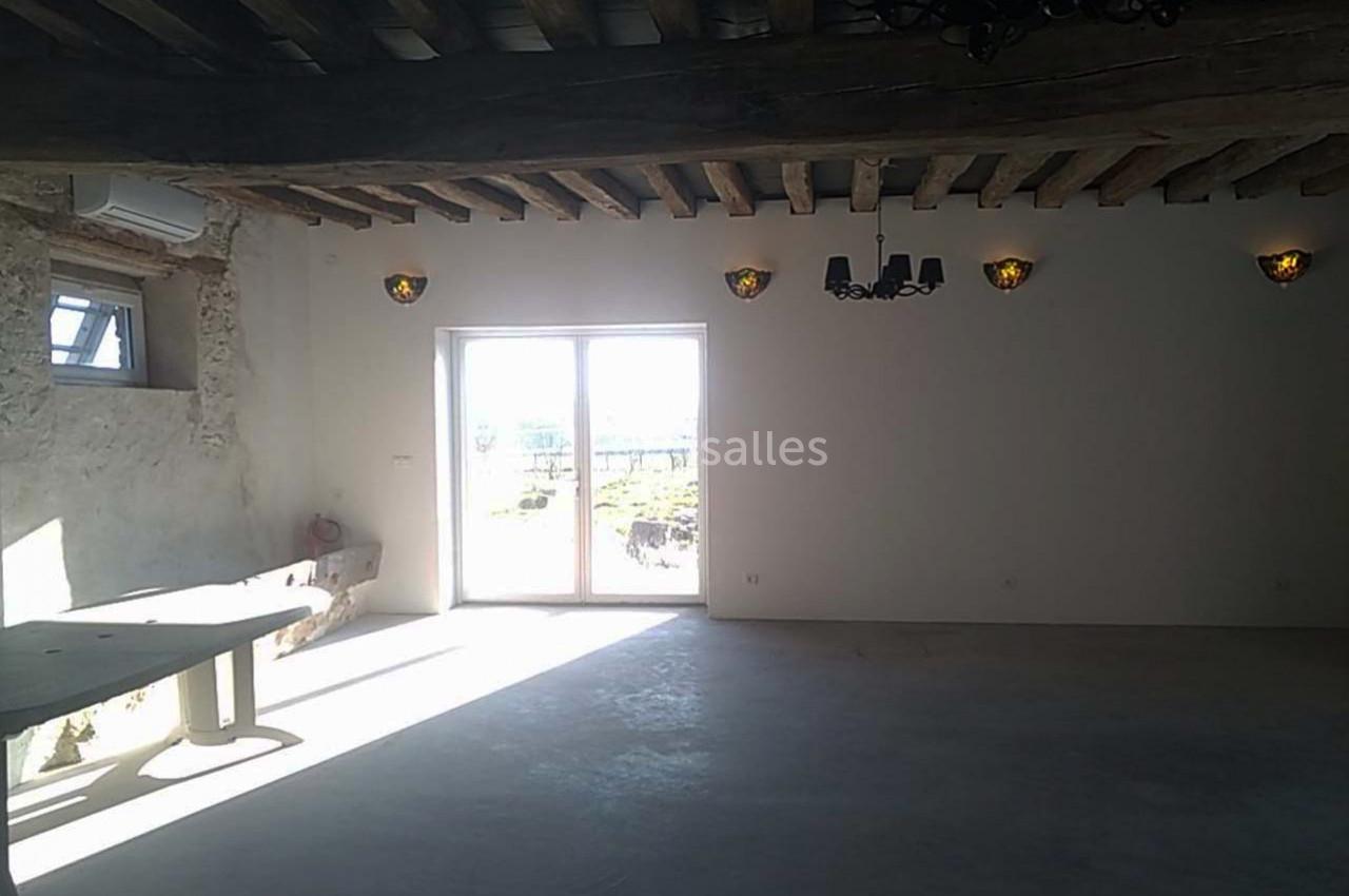 Salle lumineuse avec murs en pierre, poutres apparentes, grande baie vitrée et éclairage mural.