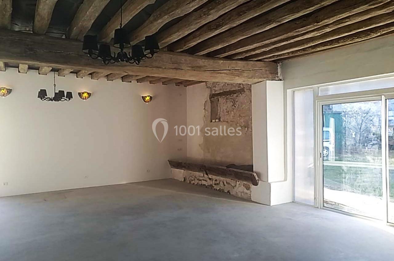 Salle lumineuse avec poutres en bois apparentes, murs clairs, sol en béton et grande baie vitrée donnant sur l'extérieur.