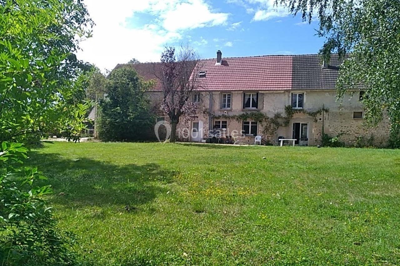 Grande maison en pierre avec toit en tuiles, entourée d'un vaste jardin verdoyant sous un ciel partiellement nuageux.