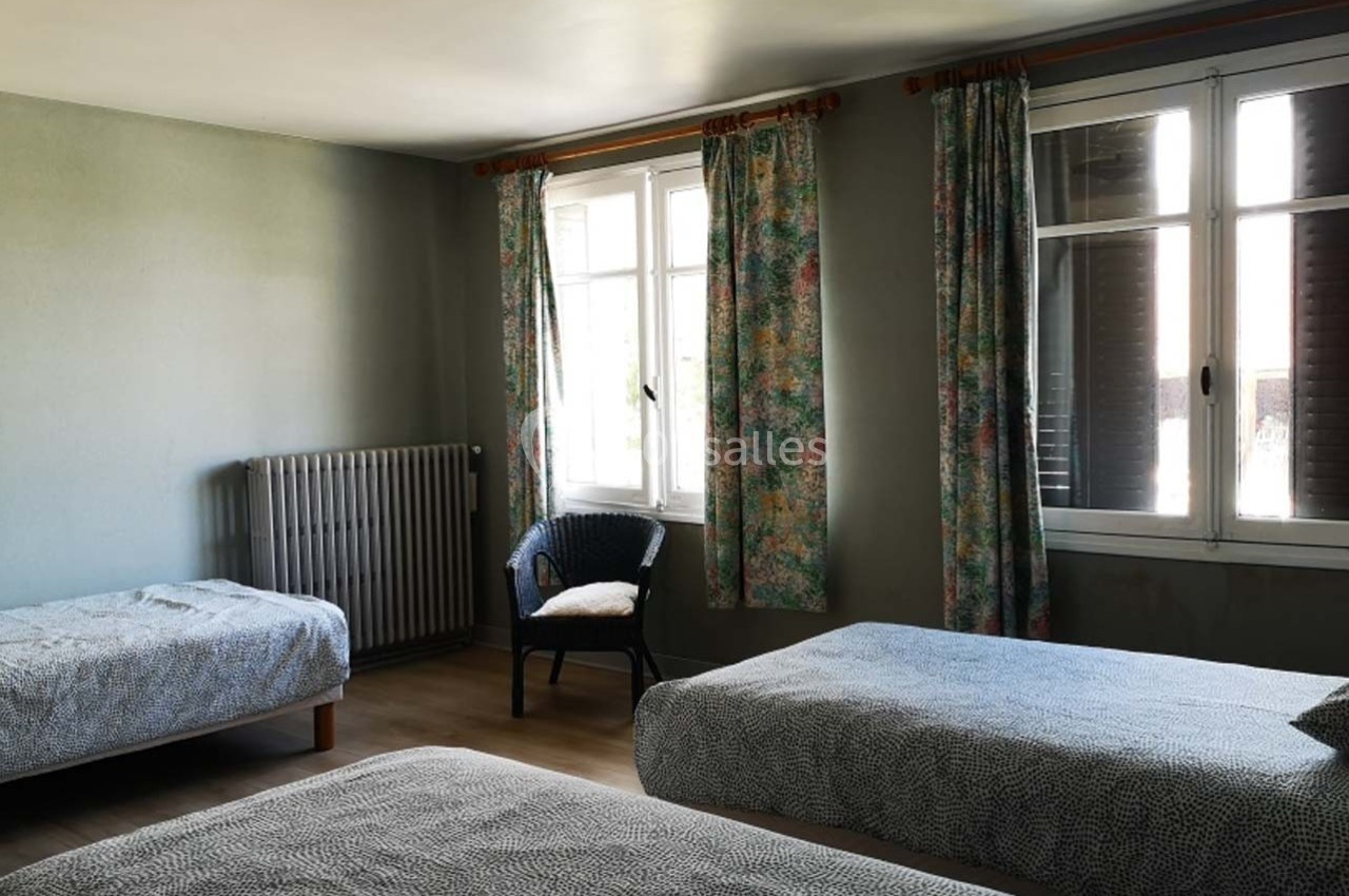 Chambre lumineuse avec trois lits simples, un fauteuil, des rideaux fleuris et un radiateur près des fenêtres.