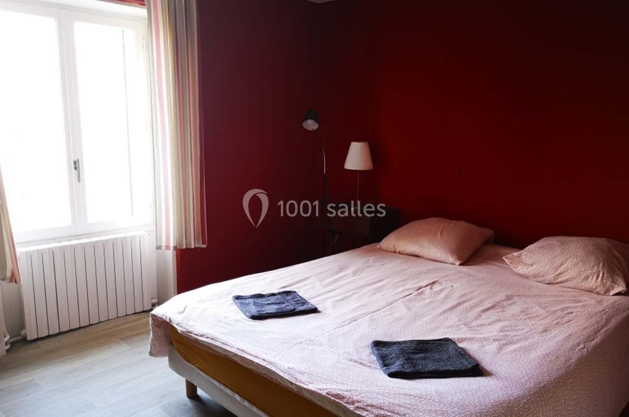 Chambre avec un lit double, draps roses, deux serviettes pliées, mur rouge, fenêtre avec rideaux clairs.