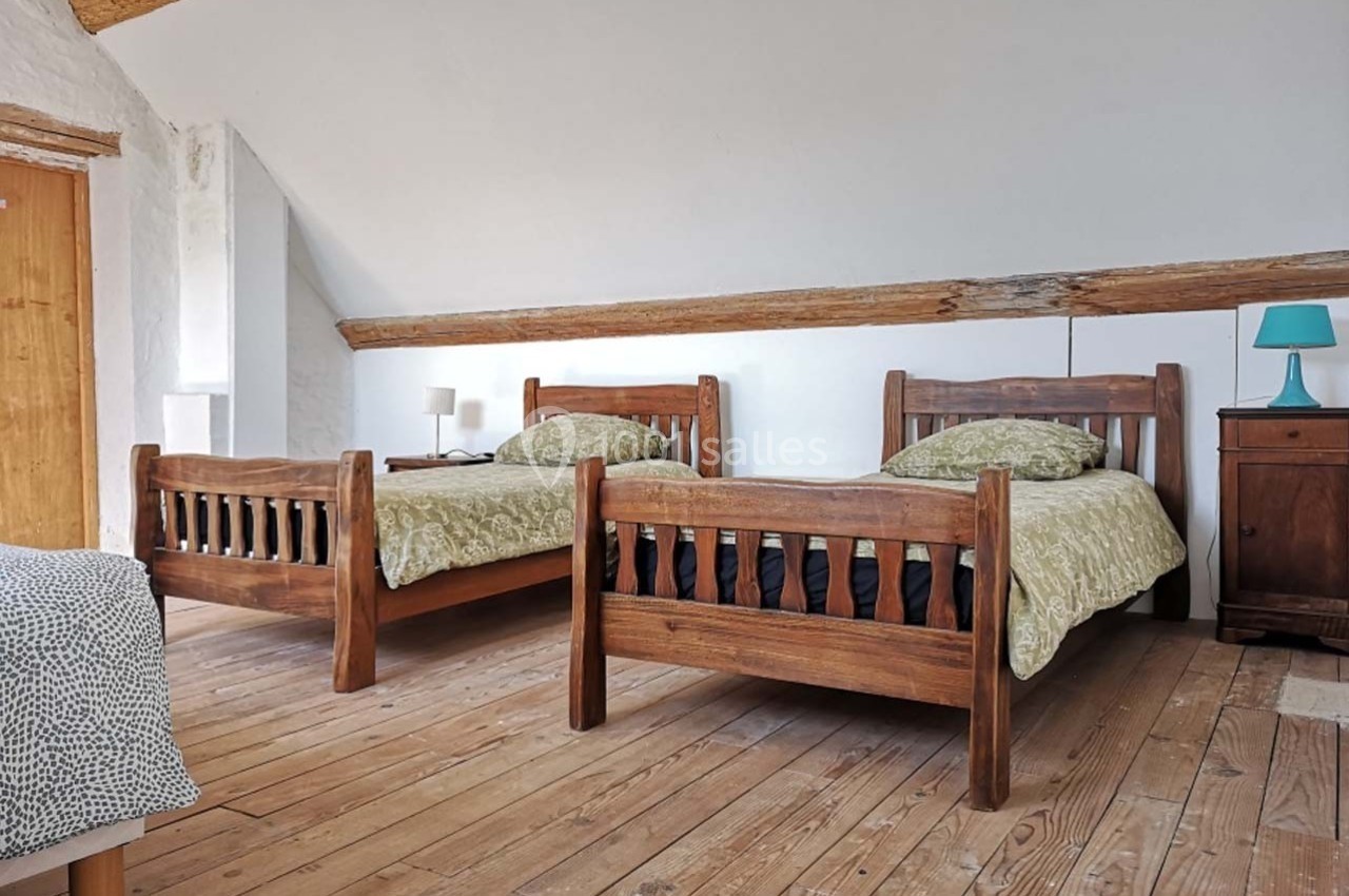 Deux lits en bois avec draps verts dans une chambre mansardée au sol en parquet, éclairée par une lampe de chevet.