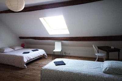 Chambre mansardée avec deux lits simples, un bureau en bois, une chaise blanche et une fenêtre de toit.
