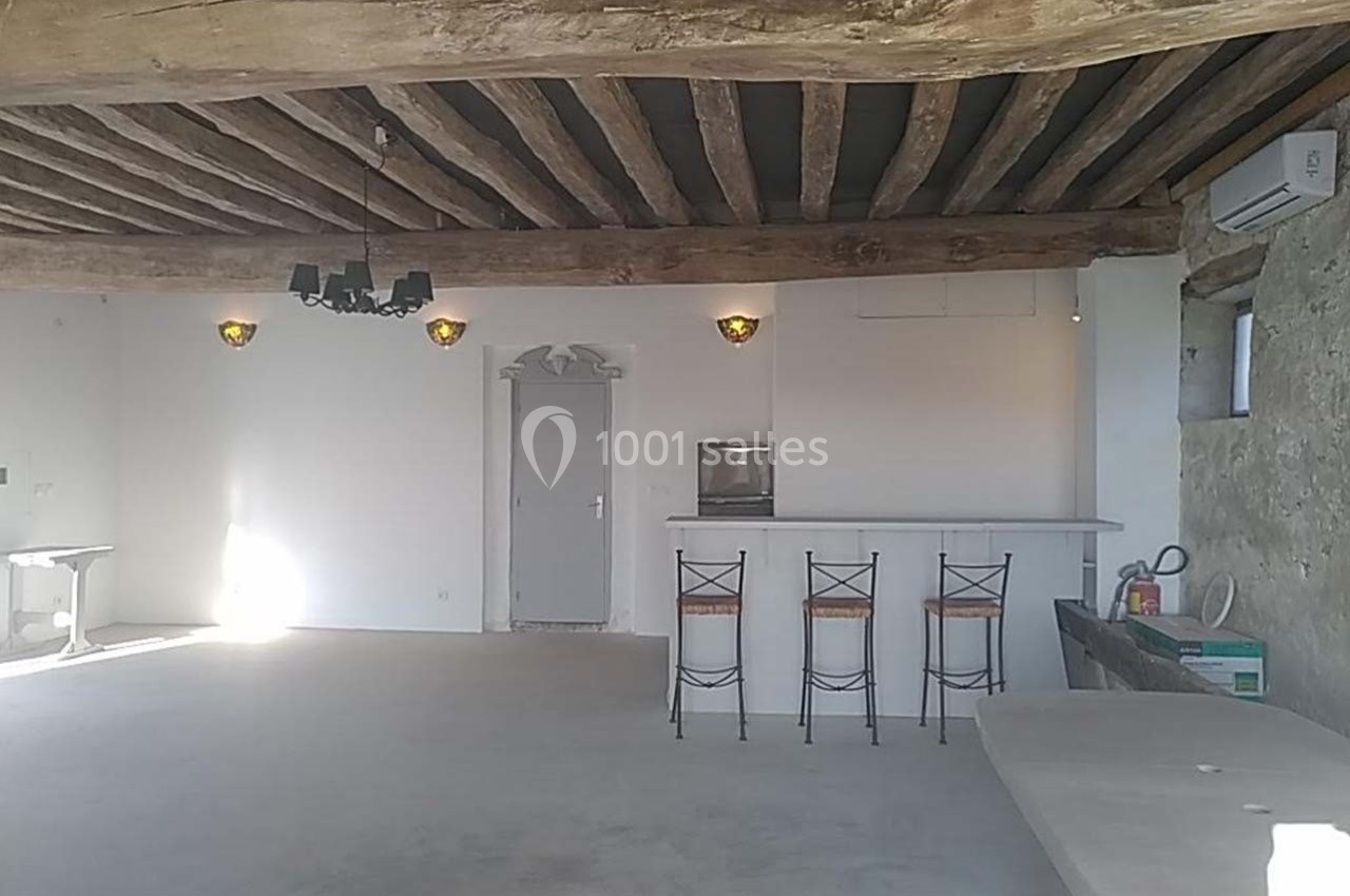 Salle lumineuse avec poutres apparentes, bar avec tabourets, murs blancs et éclairage mural.