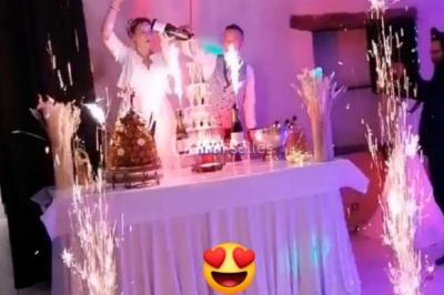 Un couple pose près d'une table décorée de fleurs et d'une pyramide de coupes de champagne dans une salle éclairée.