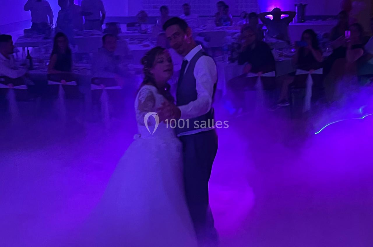 Un couple danse dans une salle éclairée par des lumières violettes, entouré de fumée et d'invités assis.