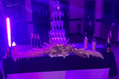 Un couple pose près d'une table décorée de fleurs et d'une pyramide de coupes de champagne dans une salle éclairée.