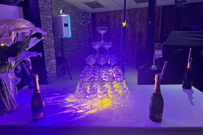 Un couple pose près d'une table décorée de fleurs et d'une pyramide de coupes de champagne dans une salle éclairée.