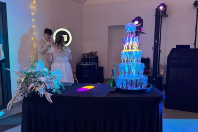 Un couple pose près d'une table décorée de fleurs et d'une pyramide de coupes de champagne dans une salle éclairée.