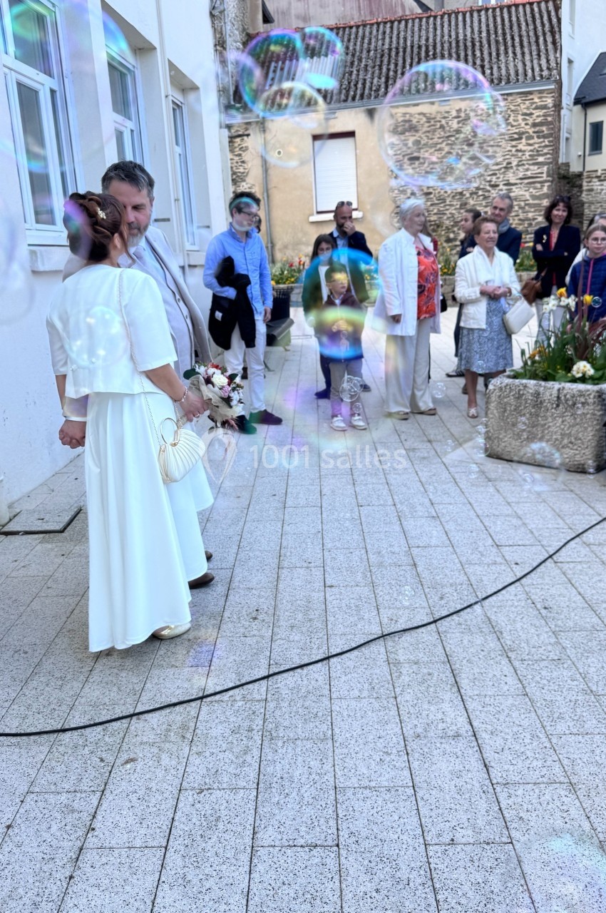Un couple en tenue de mariage se tient devant un groupe de personnes sur une place pavée, entouré de bulles de savon.