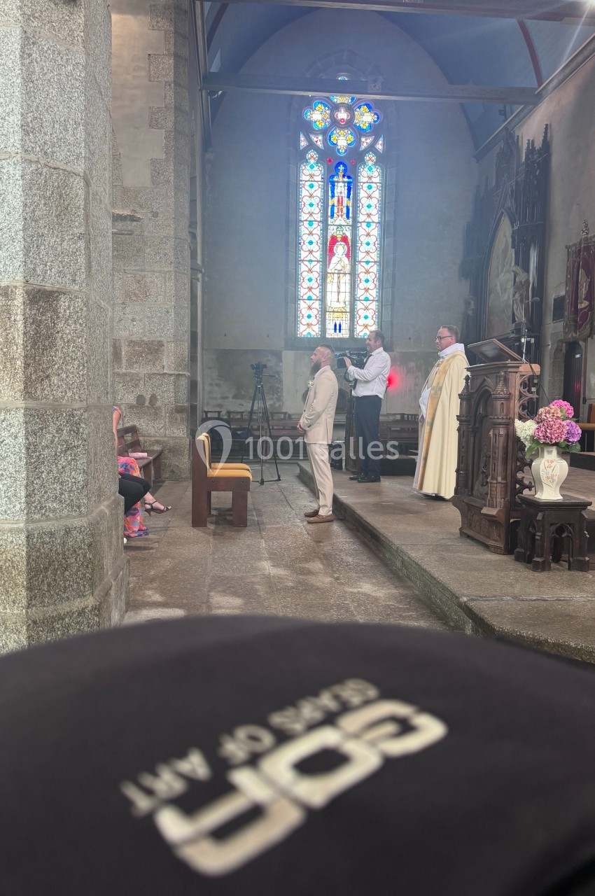 Un homme en costume est baptisé par un prêtre dans une église avec un vitrail coloré en arrière-plan.