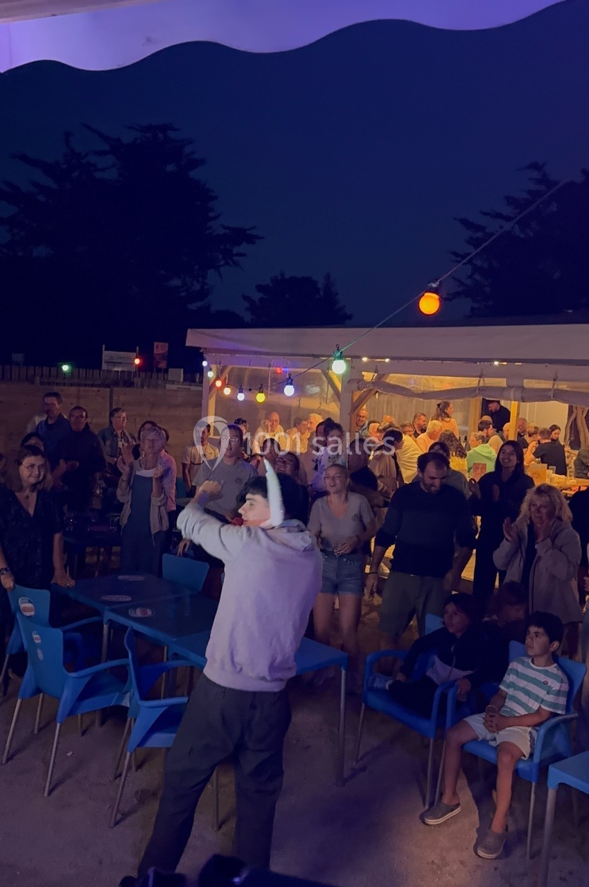 Un homme avec un micro anime une soirée en plein air devant un public assis et debout sous des guirlandes lumineuses.
