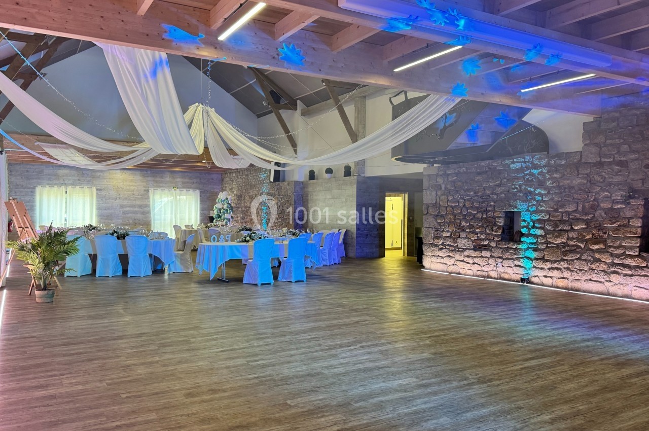 Salle de réception décorée avec des tables dressées, des nappes bleues, des voilages suspendus et un mur en pierre.