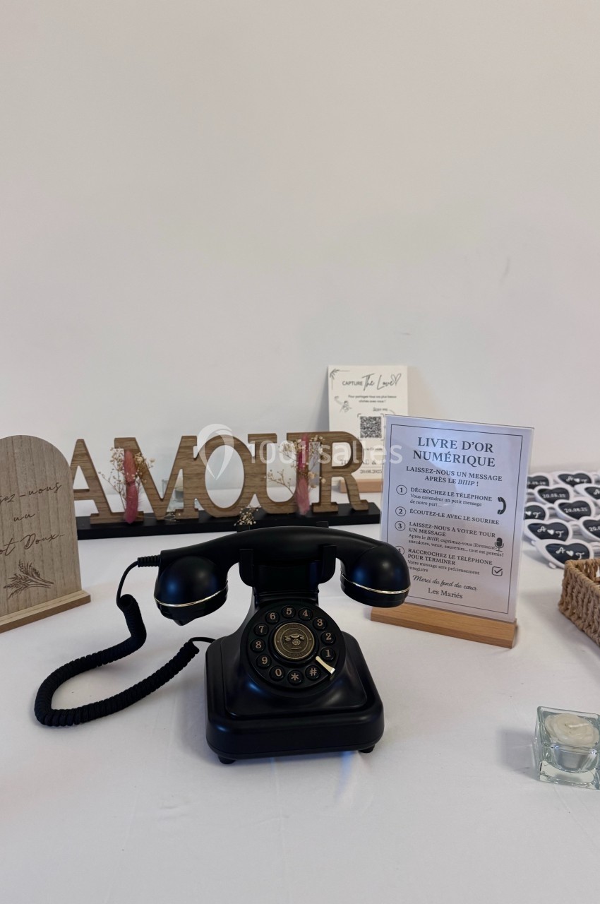 Téléphone rétro noir sur une table décorée avec des objets, dont un panneau ’AMOUR’ et un livre d'or numérique.