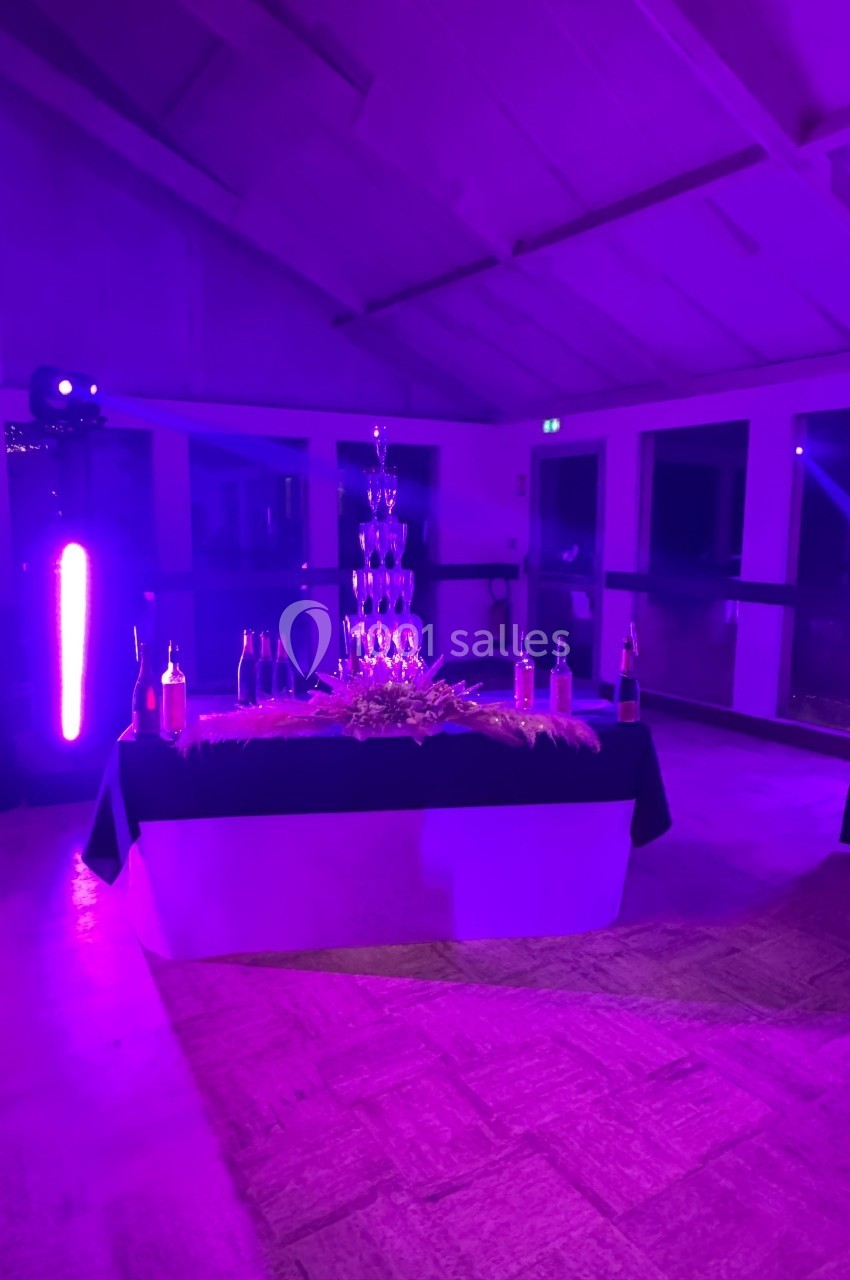 Tour de coupes en verre empilées sur une table décorée, entourée de bouteilles, éclairée par une lumière violette.