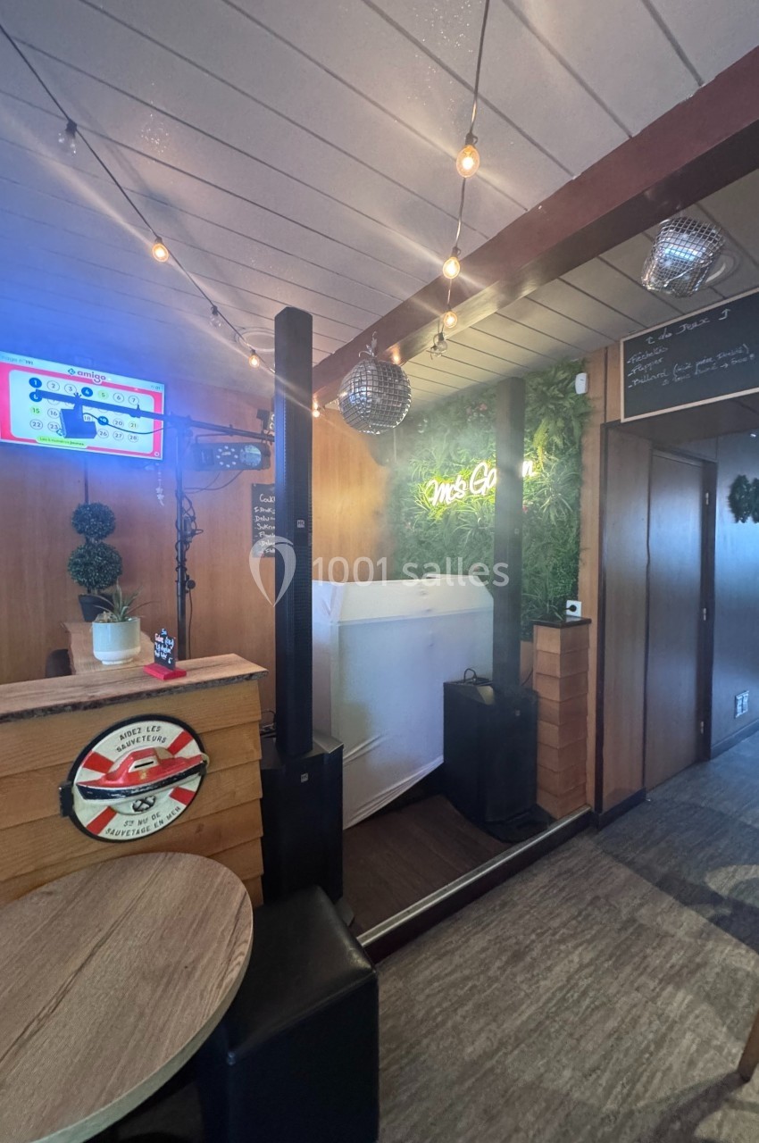 Intérieur d'un café avec décoration végétale, guirlandes lumineuses, écran mural et mobilier en bois.