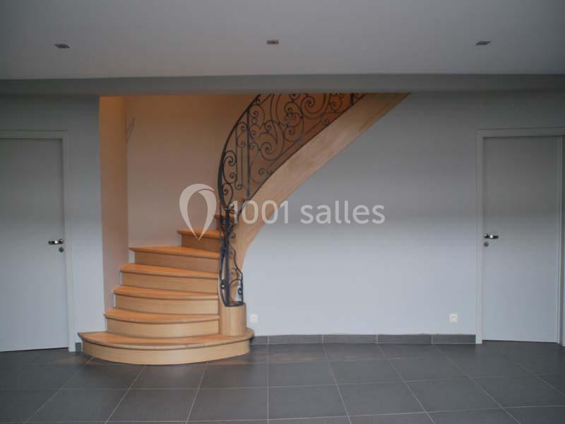 Escalier en bois avec rampe en fer forgé dans une pièce aux murs clairs et sol en carrelage gris.