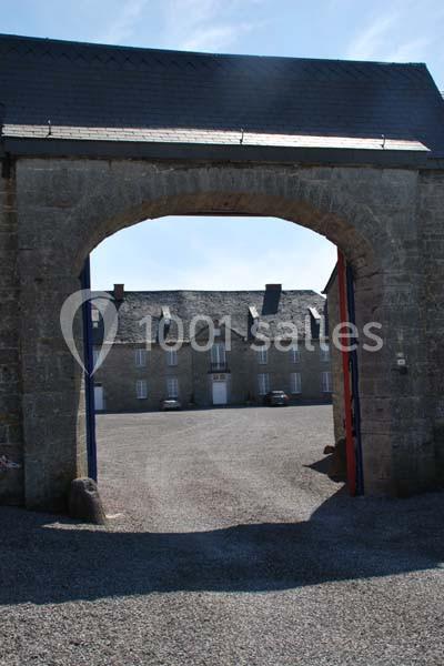 Vue d'une cour pavée entourée de bâtiments en pierre, visible à travers une grande arche en pierre.