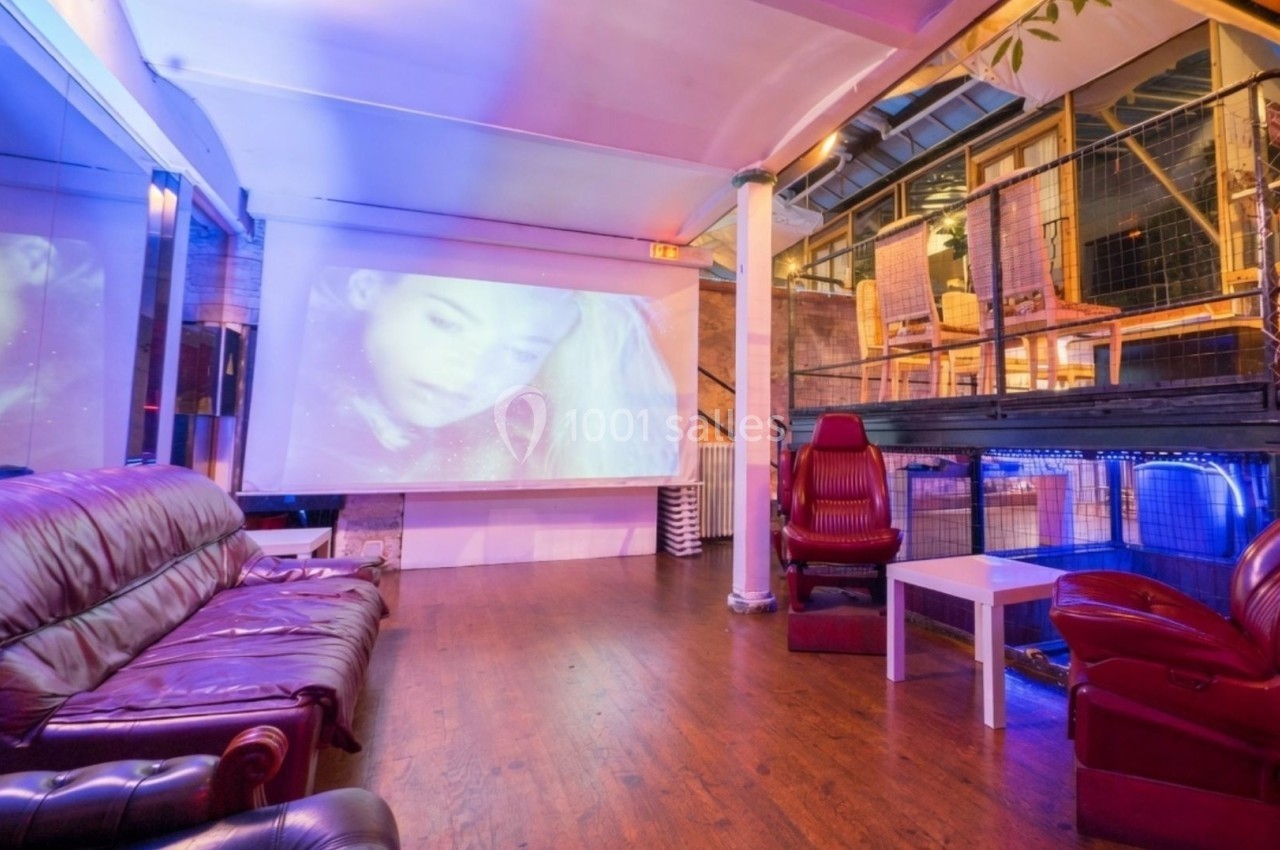 Salle avec canapés en cuir, fauteuils, écran projetant un visage, éclairage coloré et mezzanine visible en arrière-plan.