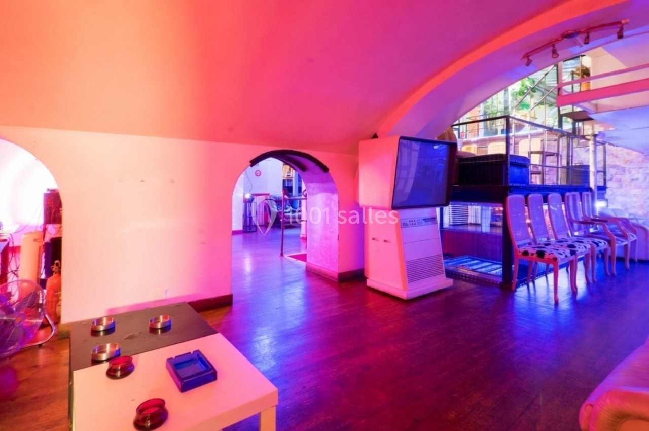Salle colorée avec éclairage rose et violet, équipée de fauteuils, tables basses et écrans, dans un espace voûté.