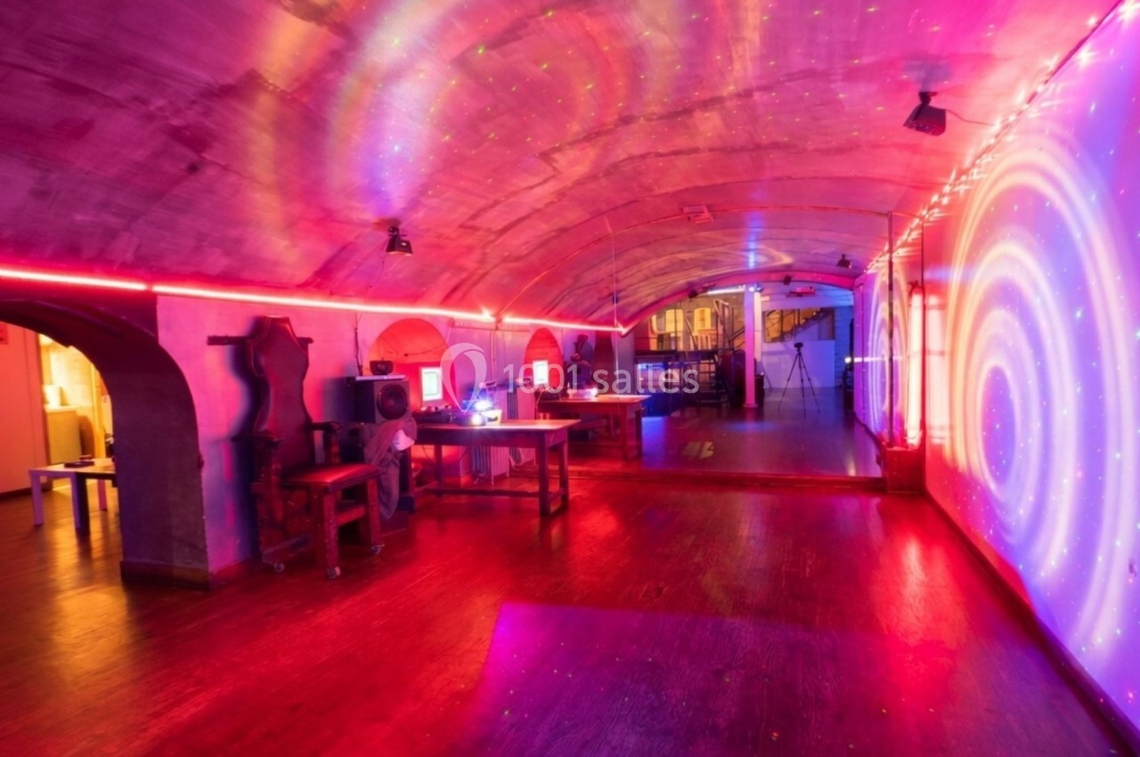 Salle voûtée éclairée par des lumières colorées, avec des bureaux, des équipements et un espace ouvert.