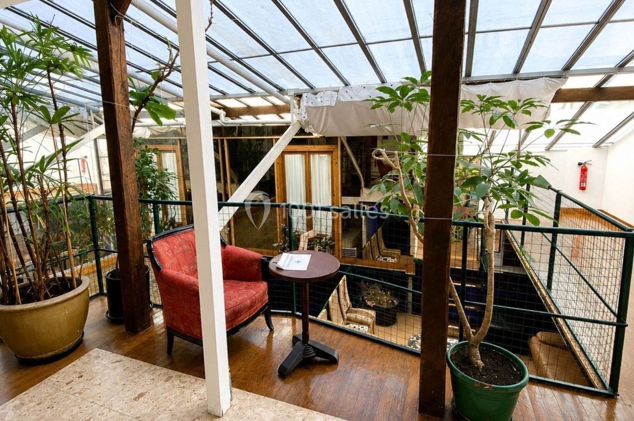Espace intérieur lumineux avec fauteuil rouge, table basse et plantes, sous une verrière en bois et métal.