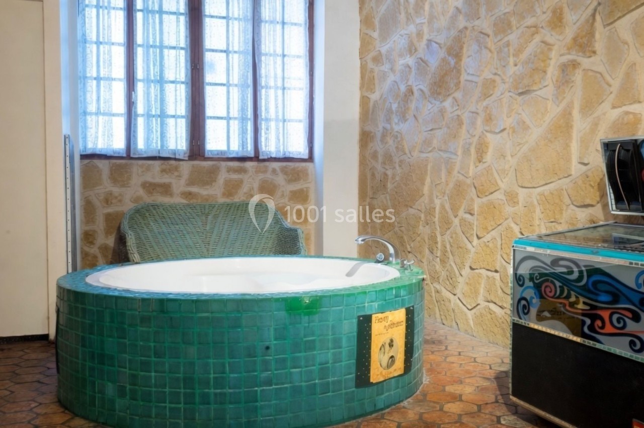 Baignoire ronde en mosaïque verte dans une pièce aux murs en pierre, avec une fenêtre et un fauteuil en arrière-plan.